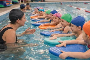Gyógyúszás foglalkozás egy uszodában: gyerekek kis csoportja úszólapokkal a vízben, miközben a gyógyúszás-oktató játékos, biztató módon vezeti a gyakorlatokat.