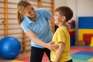 Gyógytestnevelő szakember játékos formában segít egy alsó tagozatos gyermeknek a helyes testtartás beállításában egy színes tornateremben.