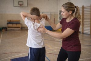 Iskoláskorú gyermek hátulról látható, karjait a feje fölé emeli egy tornateremben, miközben egy felnőtt gyógytestnevelő segíti a vállöv és a lapocka mozgását.