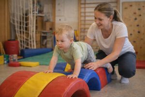 Másfél év körüli kisgyermek négykézláb mászik egy puha mozgásfejlesztő eszközön, miközben egy felnőtt támogatóan mellette van egy fejlesztőterápiás teremben.