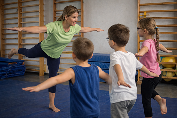 Gyógytestnevelő tanár játékos egyensúlyozó gyakorlatot vezet gyermekcsoport részére egy felszerelt tornateremben a Fővárosi Pedagógiai Szakszolgálatnál.